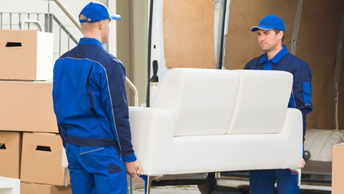 Two uniformed movers carrying a white loveseat out of a moving truck